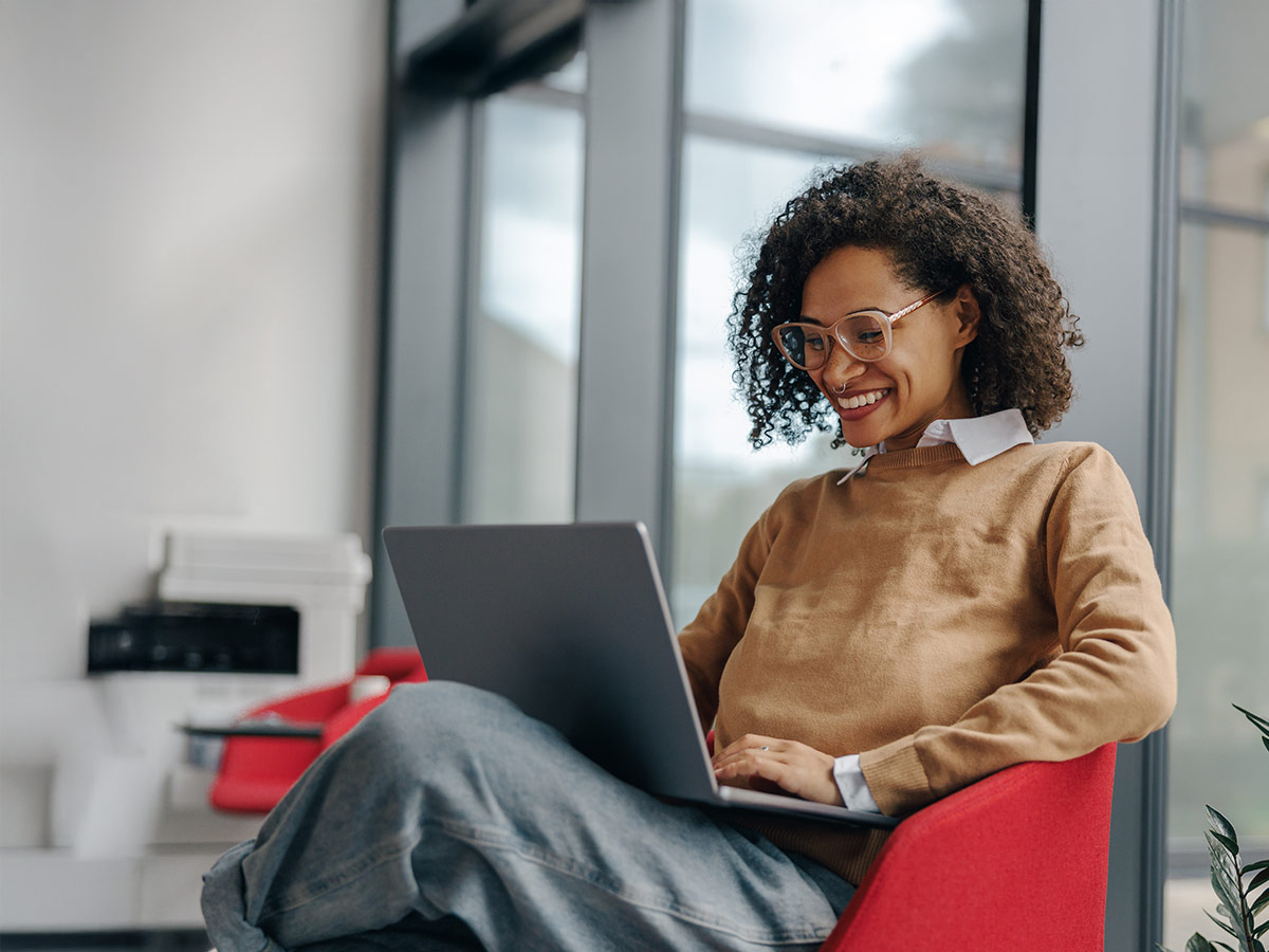 Photo of a young woman sitting in an office chair on her laptop smiling